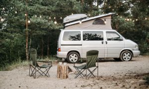Campingbus mit Dachzelt im Wald bei Lichterketten &ndash; gem&uuml;tlicher Outdoor-Stellplatz mit zwei Campingst&uuml;hlen im Sand